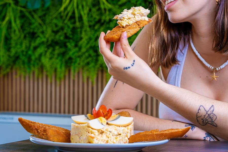 Woman outdoors taking a bite of a crostini from a plate of cheese and bread on a patio ground table.