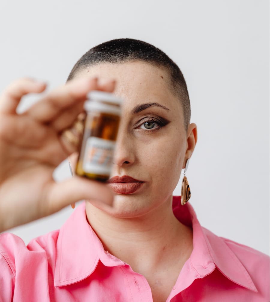 Person with a shaved head holding a brown medicine bottle toward the camera, label blurred, wearing a pink shirt.